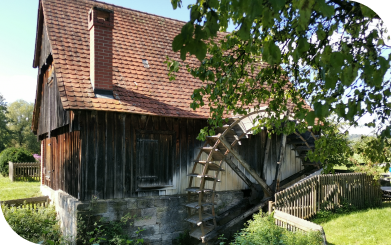 Eine Fotografie der Ölmühle von außen. Zu sehen sind das Gebäude und das Mühlrad.